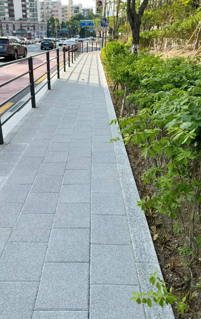 디에이치 퍼스티어 아이파크 단지 바깥 보도(세로 사진). 밝은 회색 블록 길이 난간과 화단 사이로 곧게 뻗고 멀리 도로와 건물이 보인다.
