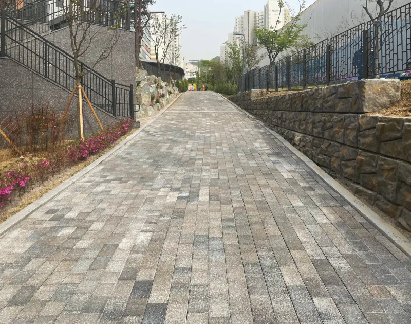 위례 자이 더 시티 단지 옆 보행로. 회색·베이지 혼합 블록 길이 완만한 오르막으로 이어지고 한쪽에 돌 무늬 옹벽과 난간이 있다.