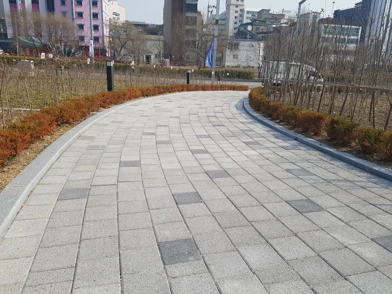 수원보훈원 곡선 산책로. 밝은 회색 블록 사이에 짙은 회색 블록이 드문드문 섞인 길이 관목 생울타리를 따라 휘어진다.