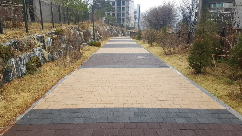 마포 현석 래미안 옹벽 아래 긴 산책로. 베이지 블록 바탕에 짙은 회색 블록 띠가 가로로 들어간 길이 마른 풀 화단을 따라 곧게 이어진다.