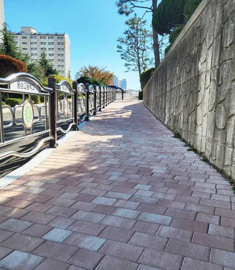 양산 웅상고등학교 옹벽 옆 경사 보행로. 무늬 옹벽과 학교 난간 사이로 분홍빛 블렌딩 블록 길이 길게 이어지며 햇빛과 그늘이 대비된다.