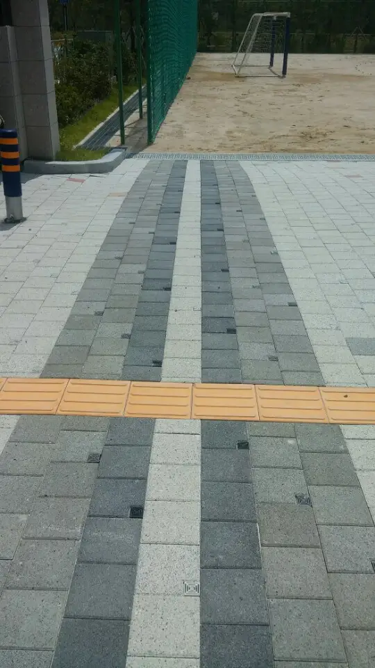위례 한빛초등학교 운동장 옆 통로 근접. 밝은 회색과 짙은 회색 블록이 세로 띠로 번갈아 깔리고 노란 점자블록 줄이 가로지른다.