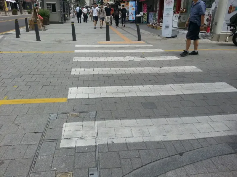 신촌 연세로 횡단 구간. 짙은 회색 블록 차로 위에 흰 블록으로 횡단보도 줄을 깔았고, 건너편 보도 입구에 볼라드와 노란 점자블록이 있다.