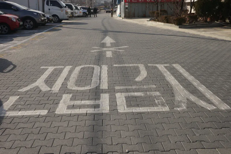 풍납동 차도블록 도로. 짙은 회색 맞물림 블록이 깔린 길 위에 흰 노면 글씨와 화살표가 그려져 있고 왼쪽에 주차 차량이 줄지어 있다.