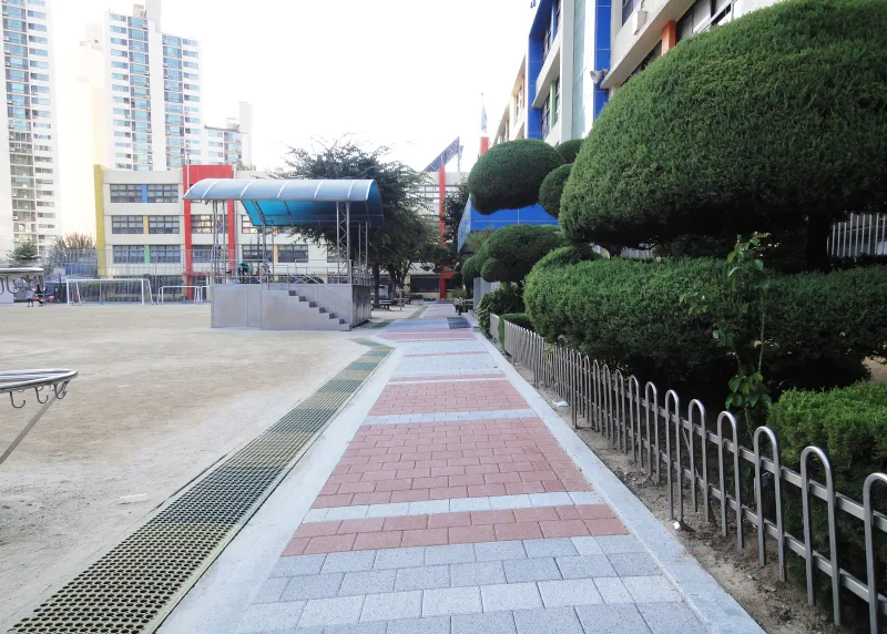 중계초등학교 향나무 화단 옆 통로. 적갈색 블록과 밝은 회색 블록 띠가 울타리와 배수 그레이팅 사이로 교사 쪽까지 이어진다.