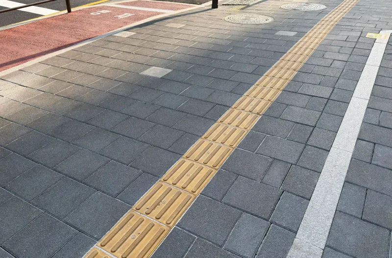 잠실 래미안 아이파크 보도 블록 근접. 짙은 회색 사각 블록 위로 노란 점자블록과 흰 선이 나란히 지나고 위쪽에 적색 포장이 보인다.