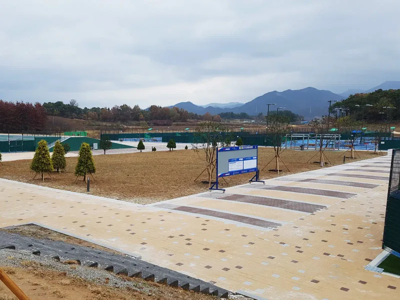 완주군 테니스장 코트 사이 산책로. 베이지 블록 길에 짙은 갈색 가로 띠가 반복되고 옆으로 어린 침엽수와 안내판, 먼 산이 보인다.