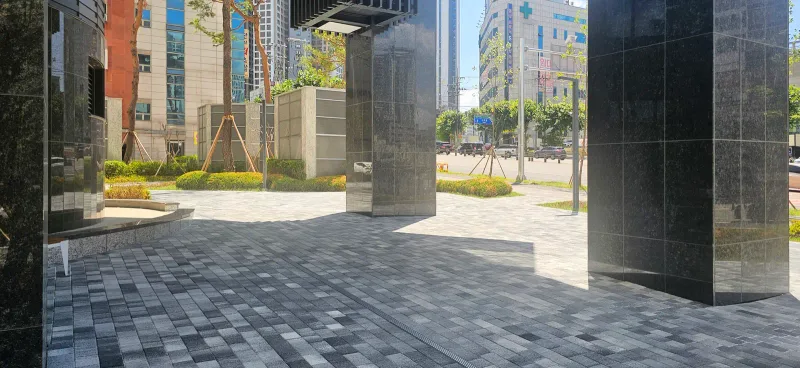 대구 달서 푸르지오 시그니처 필로티 아래 보행 공간. 검은 석재 기둥 사이로 회색 계열 혼합 블록이 넓게 깔려 있고 뒤로 화단과 도로가 보인다.