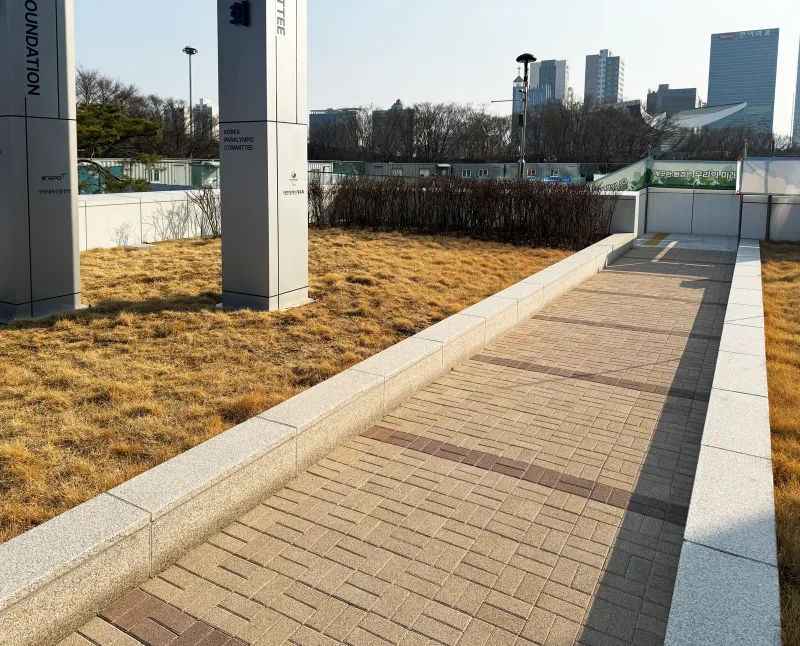 올림픽회관 기념 기둥 옆 보행로. 베이지 블록 길에 적갈색 블록 가로 띠가 들어가 있고 낮은 화강석 경계가 둘러싼다.