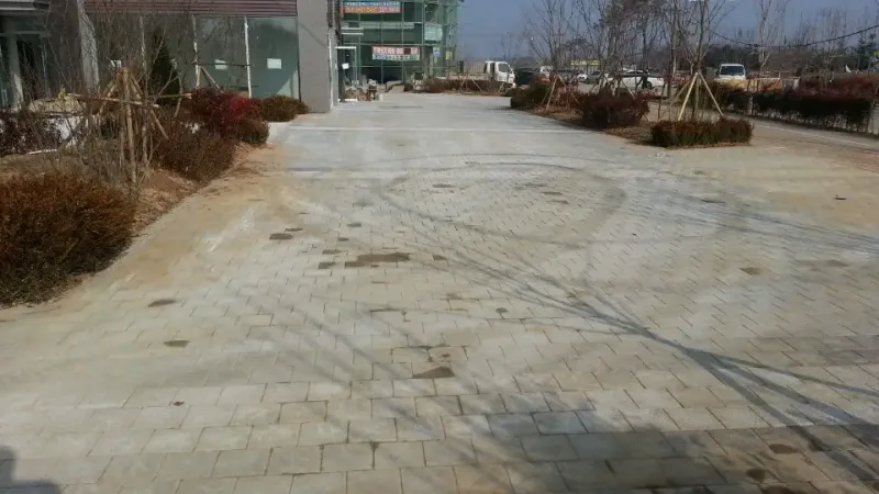 전주 우미린 단지 안 넓은 보행 광장. 밝은 회색 블록이 넓게 깔려 있고 둥근 관목 화단이 줄지어 있으며 뒤로 상가 건물이 보인다.