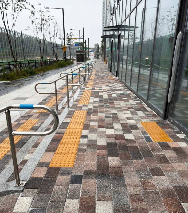 고양 풍동2지구 더샵 엘로이 경사 보도. 갈색·회색 혼합 블록 위로 노란 점자블록이 이어지고 스테인리스 손잡이 난간이 경사를 따라 설치돼 있다.