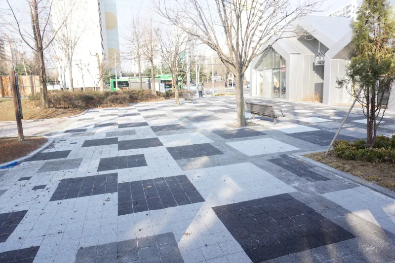 월계 한내도서관 광장을 길 쪽에서 본 모습. 흰빛 블록 사이로 짙은 회색 블록 면이 크게 박혀 있고 나무 화단과 벤치 뒤로 건물이 보인다.