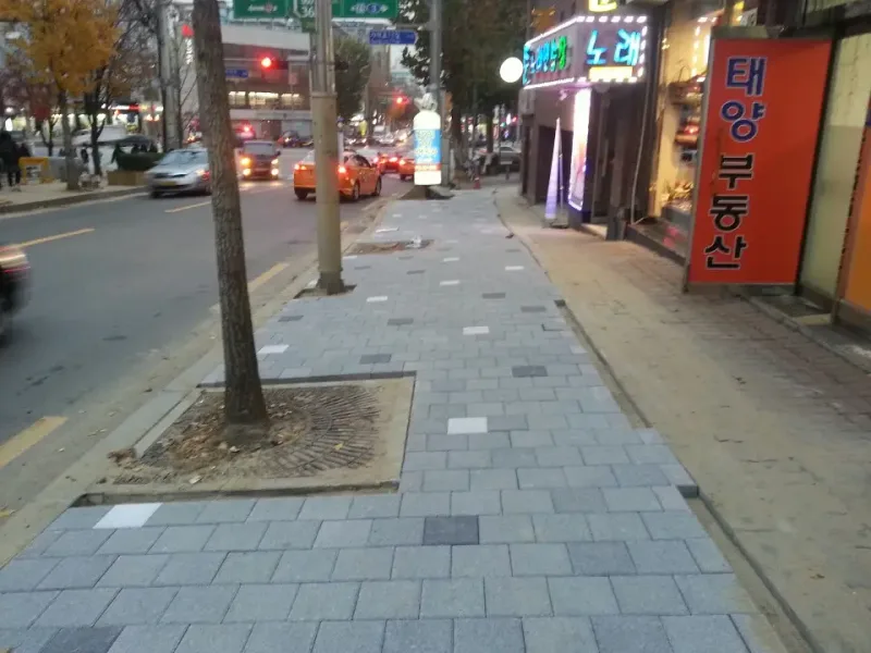 송파구 오금로 상가 앞 보도 시공 중 모습. 회색 사각 블록 사이에 흰 블록과 짙은 회색 블록이 드문드문 섞여 깔려 있고 건물 쪽 가장자리는 아직 마감 전이다.