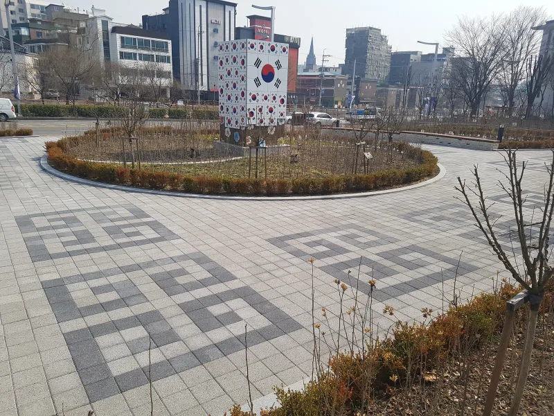 수원보훈원 광장 바닥 무늬. 짙은 회색 블록으로 꺾인 사각 문양을 짜 넣은 밝은 회색 블록 바닥 뒤로 원형 화단과 태극기 조형물이 보인다.