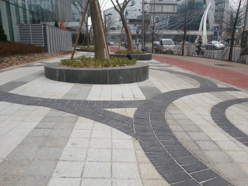 판교 SK 사옥 앞 광장. 밝은 회색 블록 바닥에 짙은 회색 블록 곡선 띠가 원형 나무 화단들을 휘감으며 이어지고, 옆으로 붉은 자전거도로가 지난다.