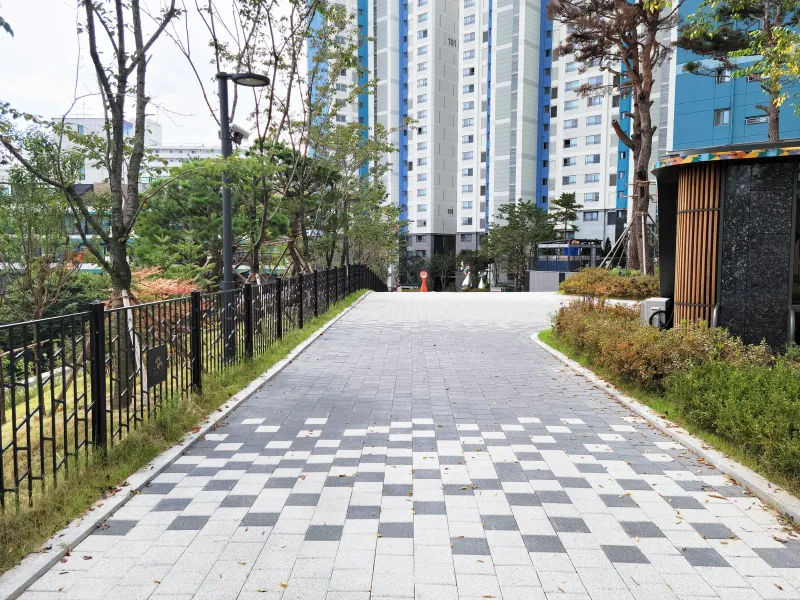 더샵 동천 이스트포레 단지 내 보행로. 회색 사각 블록 사이에 짙은 회색·흰색 블록이 체크무늬처럼 섞여 있고 양옆에 울타리와 화단, 뒤로 아파트가 서 있다.