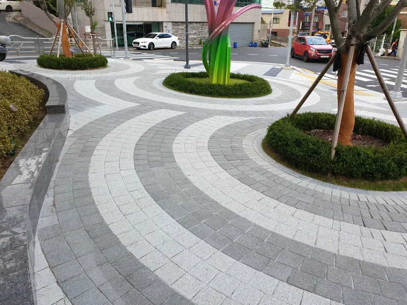e편한세상 부산항 단지 입구 광장. 연회색·짙은 회색 블록이 둥근 나무 화단을 감싸며 겹겹의 곡선 띠로 깔려 있다.