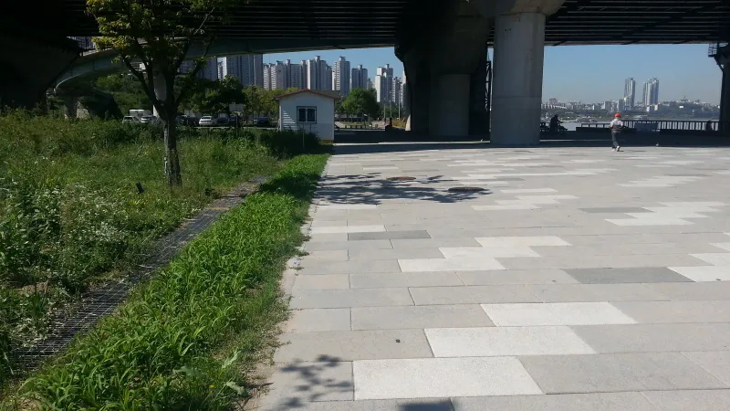 한강공원 다리 아래 광장. 밝은 회색·흰색·짙은 회색 대형 직사각 슬라브블록이 엇갈려 깔린 넓은 바닥 너머로 한강 난간과 강 건너 고층 아파트가 보인다.