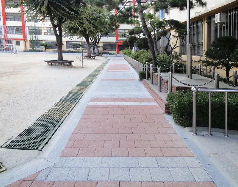 중계초등학교 교사 앞 운동장 가장자리 길. 적갈색 블록 구간과 회색 블록 띠가 번갈아 깔린 길 옆으로 긴 배수 그레이팅과 난간이 있다.