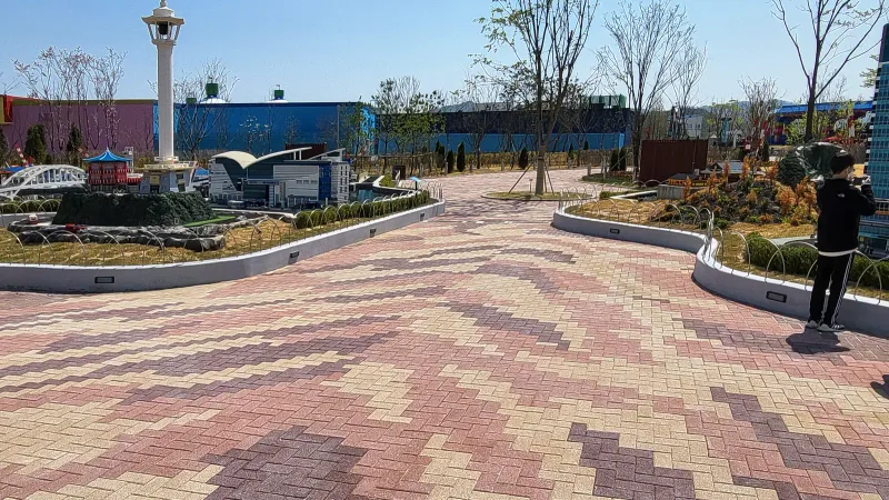 춘천 레고랜드 미니랜드 갈림길. 분홍·베이지·자주색 블록이 헤링본으로 깔려 굵은 물결무늬를 만들고 양옆 곡선 화단 안에 축소 모형이 놓여 있다.