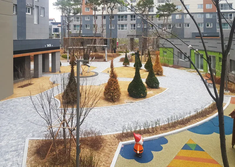 동탄2 아이파크 단지 안 놀이터 주변. 회색 블록 산책로가 곡선으로 휘어 돌고 원뿔형 나무 화단과 아파트 동이 둘러싸고 있다.