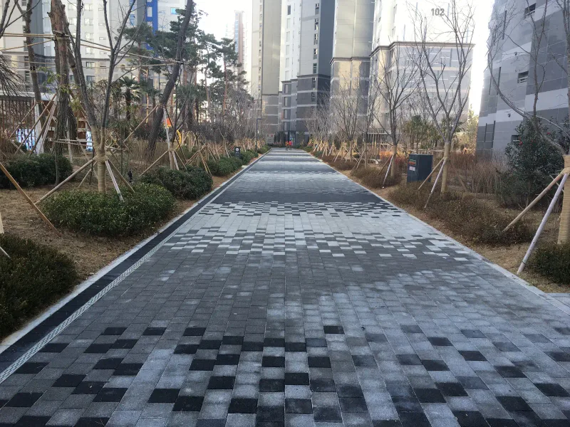 창원 용호 더샵 단지 내 곧은 보행로. 짙은 회색과 밝은 회색 블록이 모자이크처럼 섞이며 앞쪽으로 갈수록 짙어지고 양옆에 관목 화단이 있다.