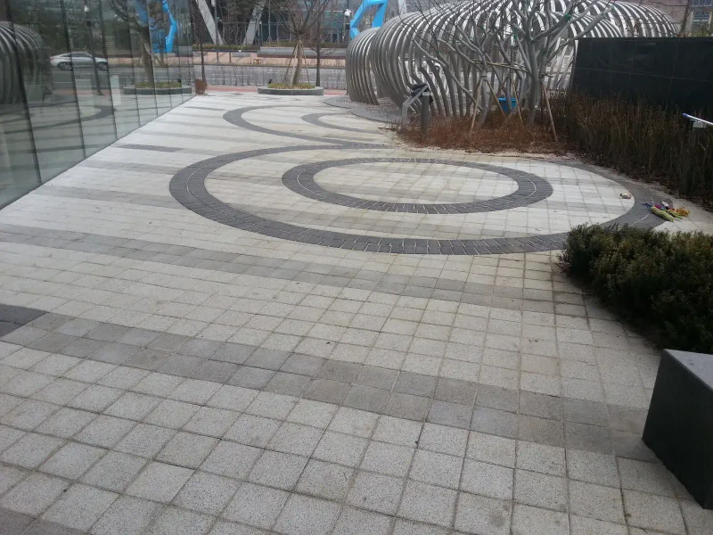 판교 SK 사옥 앞 휴게 공간. 밝은 회색 블록 바닥에 짙은 회색 블록으로 겹겹이 동심원을 그렸고, 뒤로 곡선형 조형 구조물과 화단이 보인다.