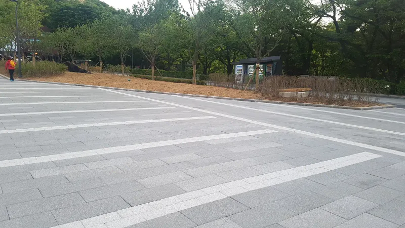 유스트림 광장의 넓은 포장면. 회색 블록 바탕에 흰빛 블록 띠가 사선으로 교차하며 뒤편 화단과 안내판 쪽으로 이어진다.