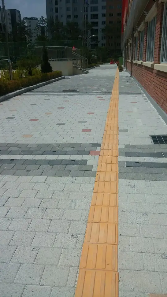 위례 한빛초등학교 교사동 옆 보행로. 밝은 회색 블록 길 가운데로 노란 점자블록이 곧게 뻗고 짙은 회색 가로 띠가 들어가 있다.