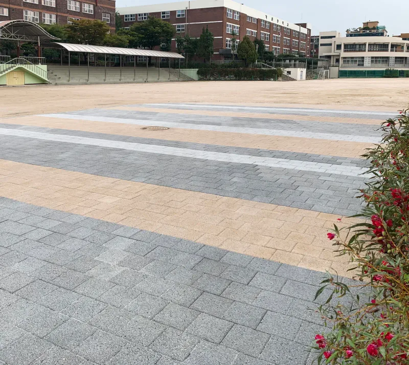 평화초등학교 운동장 앞 블록 포장 구간. 회색·베이지·흰색 블록이 넓은 가로 띠 무늬로 깔렸고 뒤로 스탠드와 벽돌 교사가 보인다.