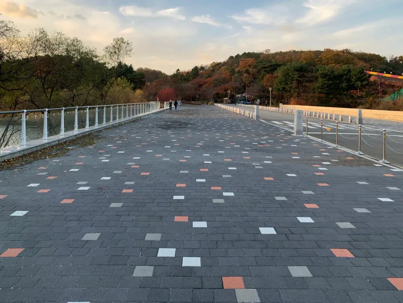 서울대공원 호숫가 순환도로의 호수 옆 넓은 보행로. 짙은 회색 블록 사이에 흰색·회색·주황색 블록이 흩어져 깔려 있고 왼쪽으로 유리 난간이 이어진다.