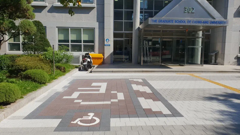 중앙대학교 대학원 건물 입구 주차면. 적갈색·흰색·짙은 회색 블록으로 글자와 휠체어 표시를 짜 넣은 장애인 주차 구역이다.