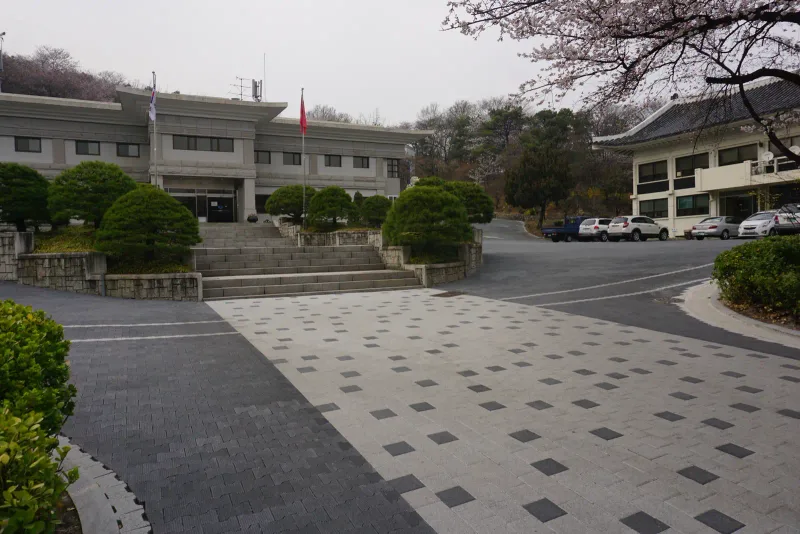 국립서울현충원 청사 앞마당. 짙은 회색 정사각 블록이 점점이 박힌 연회색 보행 광장이 짙은 회색 차도블록 진입로와 맞닿고 뒤로 청사 건물이 보인다.