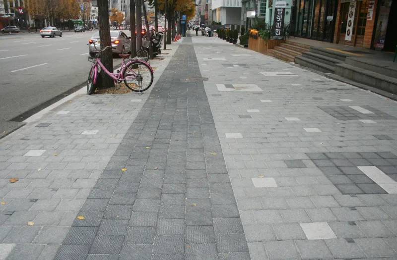 도산대로 가로수 보도. 가운데로 빗살 질감의 짙은 회색 블록 띠가 곧게 뻗고, 양옆 밝은 회색 블록 바닥에 짙은 회색·흰색 블록 무늬가 드문드문 놓여 있다.
