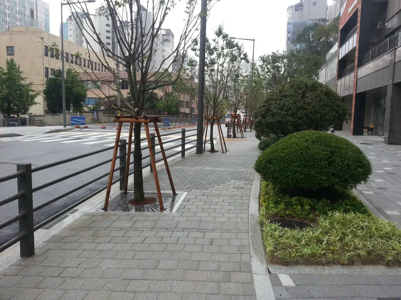 아현동 현장 도로변 보도. 회색 직사각형 블록이 깔린 보도 옆으로 새로 심은 가로수와 둥근 관목 화단이 있고, 난간 너머로 차도가 이어진다.
