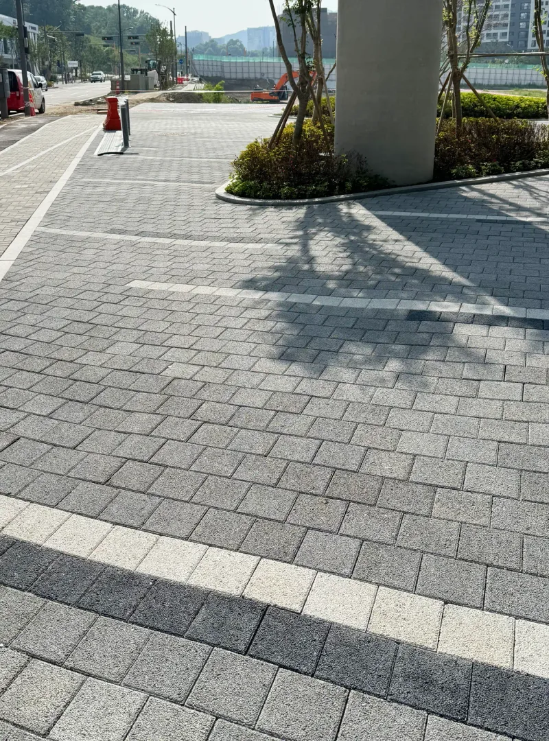 과천 펜타원 건물 기둥 옆 포장. 회색 블록 바닥에 밝은 회색·짙은 회색 가로 띠가 겹쳐 들어가 있고 나무 그림자가 드리운다.