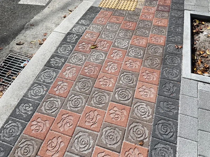 부천 장미특화거리 빗물받이 옆 보도. 분홍·적갈색 장미 무늬 블록 띠 한쪽에 배수 덮개가, 다른 쪽에 가로수 화단이 있다.