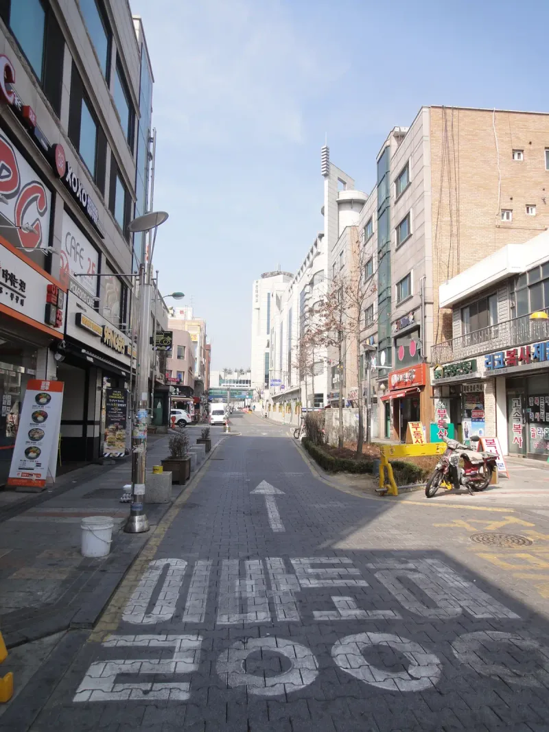 부천 차도블록 구간 내리막 거리. 블록 포장 차도 위에 흰 직진 화살표 표시가 있고 양쪽 상가 사이로 멀리 행인이 지나간다.
