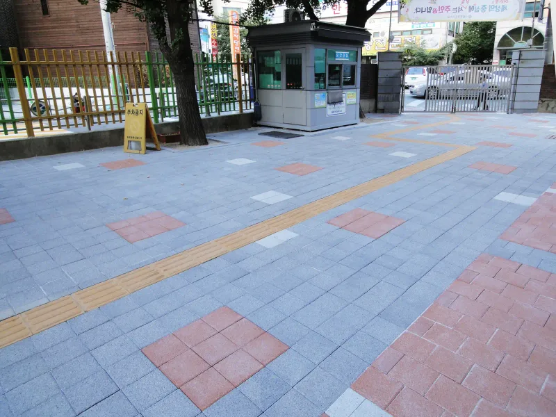 중계초등학교 정문 경비실 앞 바닥. 회색 블록 위에 적갈색 블록 사각 무늬가 드문드문 놓이고 노란 점자블록이 사선으로 이어진다.