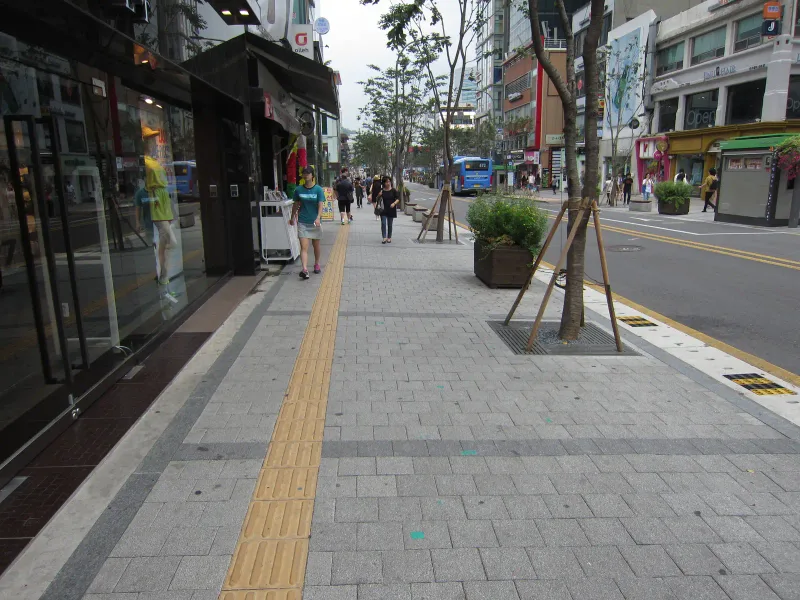 신촌 연세로 상가 앞 보도. 회색 블록 보도를 따라 노란 점자블록이 길게 이어지고, 가로수 보호판과 대형 화분 너머로 차로와 버스가 보인다.