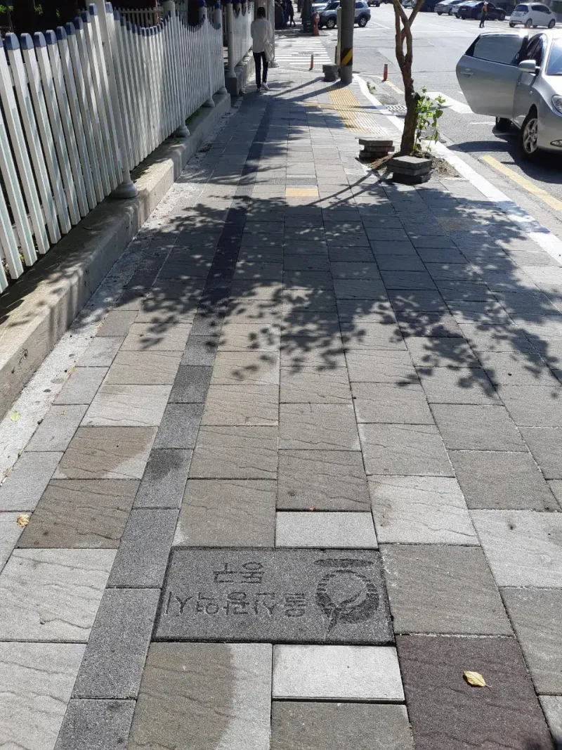 울산 낭만의거리 울타리 옆 보도. 나무 그늘이 드리운 회색·베이지·적갈색 사암 질감 블록 길에 구 상징 표지석과 노란 점자블록이 보인다.