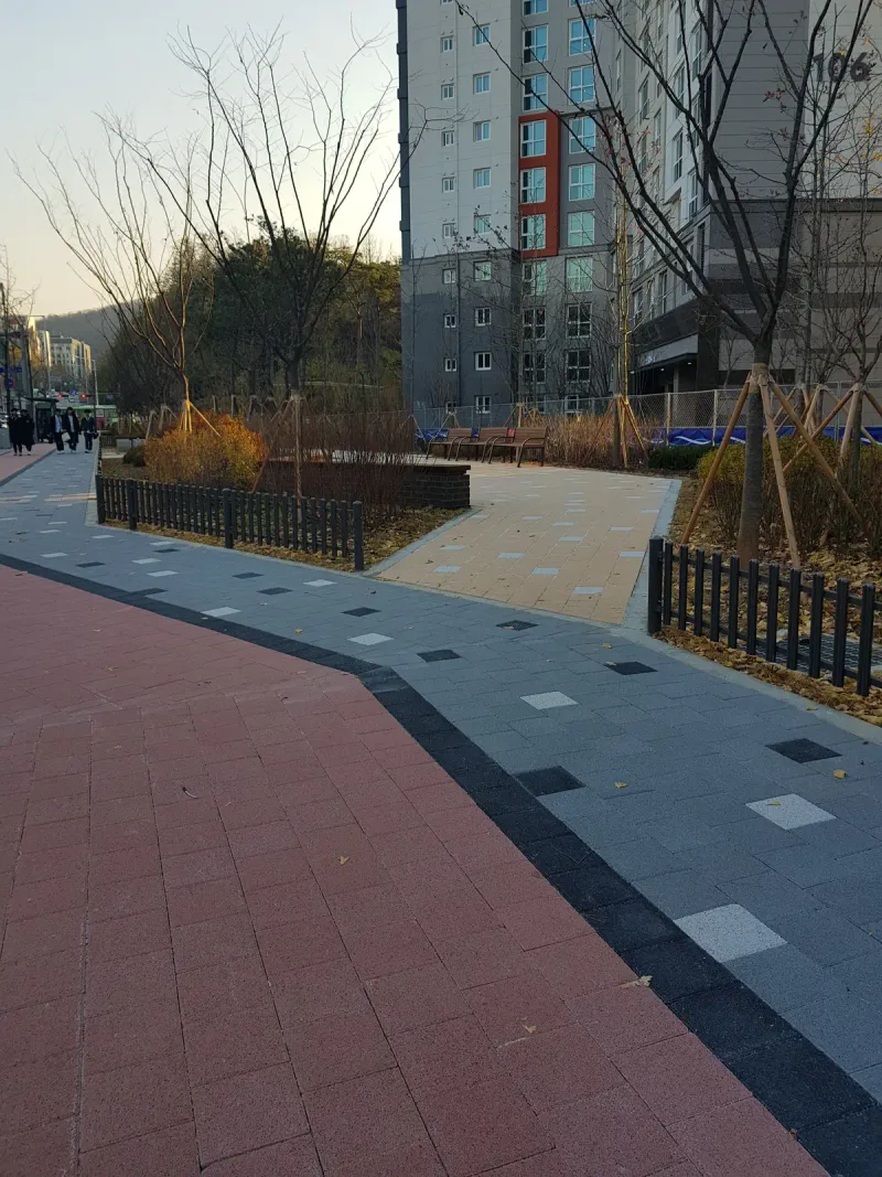 일원동 래미안 루체하임 단지 외곽 보도. 적갈색 블록 길과 짙은 회색 블록 보도가 나란히 놓이고 안쪽으로 베이지 톤 산책로가 갈라진다.