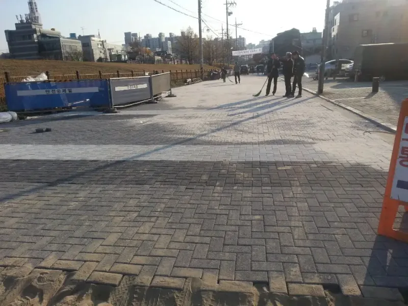 송파 저류기층 시범구간, 시공 중. 짙은 회색 블록이 헤링본 무늬로 깔린 구간과 밝은 회색 블록 띠가 이어지고, 뒤로 안전 펜스와 둑길이 보인다.