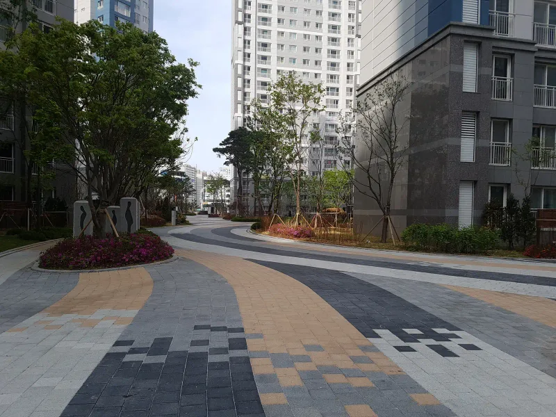 시흥 배곧 한신휴플러스 단지 곡선 보행로. 베이지·짙은 회색·밝은 회색 블록이 나란한 리본 띠로 휘어 가고 앞쪽에서 모자이크처럼 흩어진다.