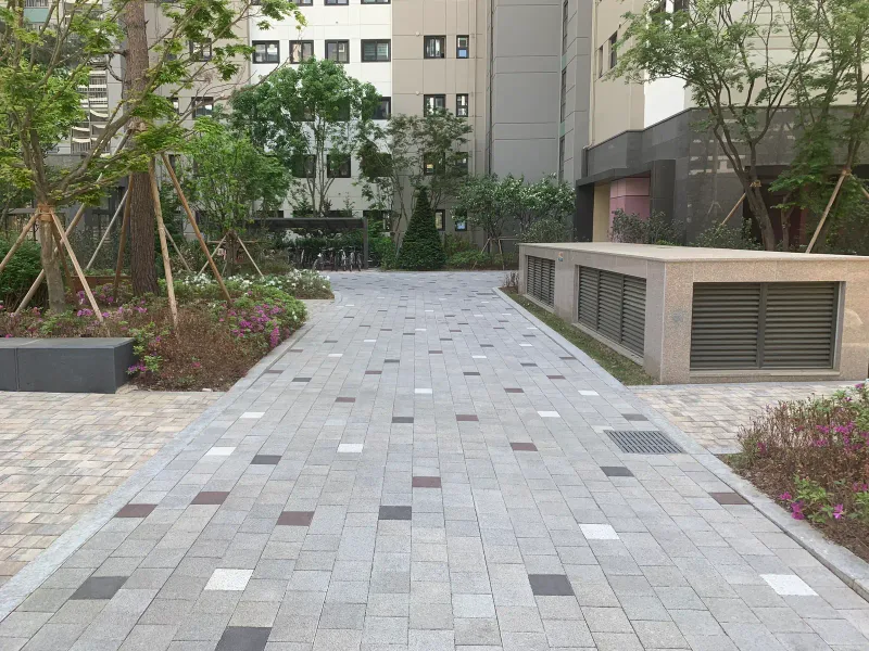 송파 헬리오시티 환기구 옆 광장형 보행로. 연회색 블록 바닥에 짙은 회색·흰색·적갈색 블록이 흩어진 무늬로 깔려 있다.