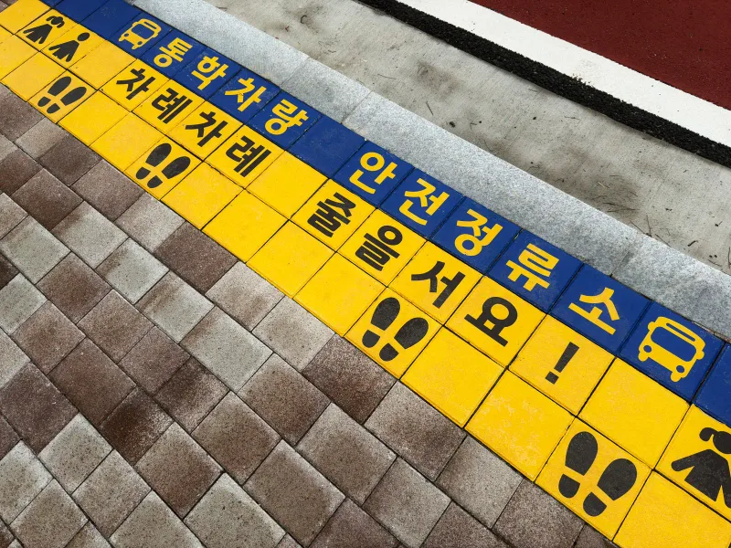 동탄2 숨마데시앙 통학차량 정류소 바닥. 갈색 혼합 블록 사이에 파란색·노란색 안내 블록이 띠를 이루고 발자국과 '줄을 서요' 글자가 새겨져 있다.