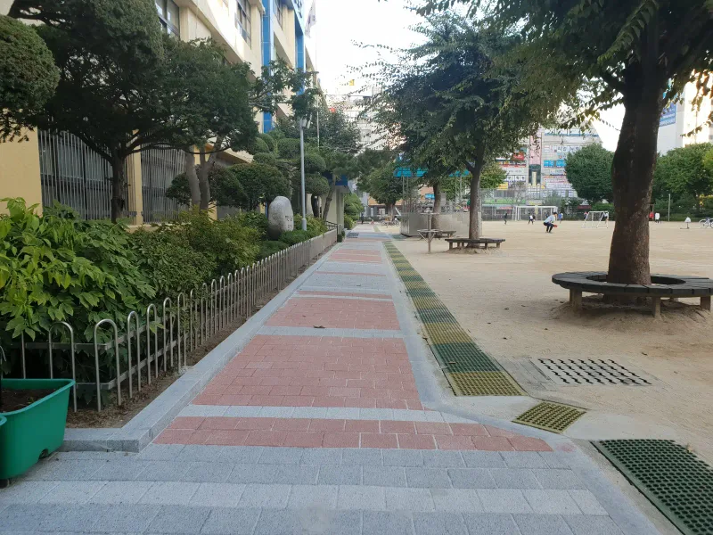 중계초등학교 운동장 옆 통로. 밝은 회색 블록 길에 적갈색 블록 띠가 반복되고 옆으로 배수 그레이팅, 나무 둘레 벤치, 낮은 울타리가 이어진다.