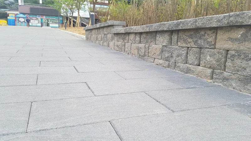 서울대공원 느티나무길 석재 화단 벽 옆. 회색 직사각형 대형 블록이 벽을 따라 비스듬히 깔려 있고 멀리 공원 건물이 보인다.