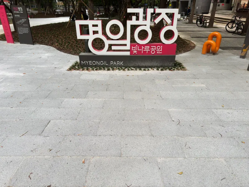 명일광장 이름 조형물 앞 바닥. 밝은 회색 긴 판형 블록이 어긋나게 깔려 있고 뒤로 화단과 자전거 거치대가 보인다.