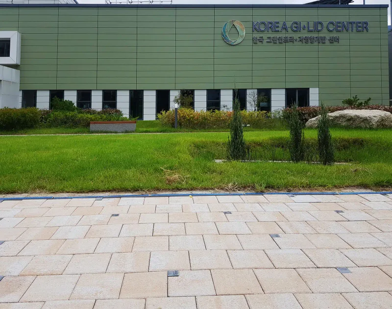 한국 GI·LID 센터 정면 보행로. 베이지·살구색 직사각형 블록이 파란 경계재를 따라 깔려 있고 뒤로 잔디밭과 센터 건물이 보인다.